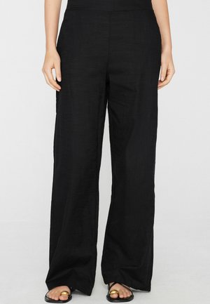 Weit geschnittene schwarze Hose, getragen mit schwarzen Sandalen mit goldenen Zehenringen, gezeigt an einer stehenden Person vor einem einfarbig hellen Hintergrund.