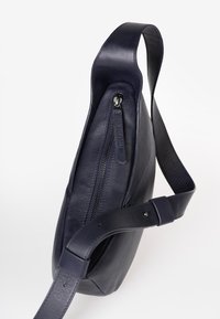 Borsa a spalla in pelle blu navy con design curvo e chiusura con zip visibile. Dotata di una tracolla piatta e superficie liscia e texturizzata.