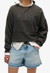 Ciemnoszara bluza z wypukłym napisem "SUPERDRY ATHLETIC" w ciemnych literach, zestawiona z jasnoniebieskimi szortami dżinsowymi o postrzępionych krawędziach.