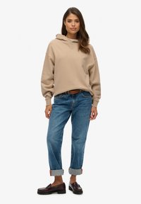 Haut à capuche beige avec une coupe décontractée, associé à un jean bleu avec les poignets retroussés et des mocassins marron foncé, avec une texture lisse et des accents minimaux.
