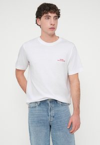 Λευκό βαμβακερό μπλουζάκι με κοντά μανίκια, με μικρό κόκκινο κείμενο στην αριστερή πλευρά του στήθους. Συνδυασμένο με τζιν παντελόνι ανοιχτού μπλε χρώματος.