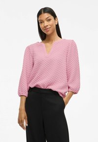 Kvinna med långt mörkt hår, klädd i en texturerad rosa blus med 3/4-ärmar och svarta högmidjade byxor, står med en hand i fickan.