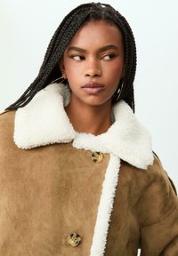 Giovane con lunghe trecce che indossa orecchini a cerchio dorati e un cappotto in shearling beige con grandi bottoni e un colletto bianco soffice.