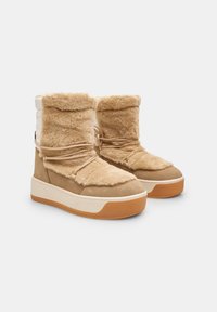 Botas de tobillo forradas de piel de color marrón claro, con una parte superior en crema y blanco, que presentan un diseño de cordones y una suela de goma con una entresuela de color marrón contrastante.
