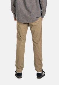 Persona vistiendo pantalones beige, zapatillas Adidas negras con rayas blancas y una camiseta gris de manga larga, vista desde atrás.