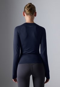 Navy dugi rukav sportski top ima prilagođeni kroj i glatku teksturu, s okruglim izrezom i diskretnim logotipom na leđima.