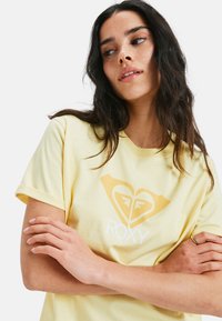 Gelbes Baumwoll-T-Shirt mit aufgerollten Ärmeln, das ein auffälliges Herzlogo und den weißen Schriftzug "ROXY" zeigt. Weiche Textur und lässige Passform.