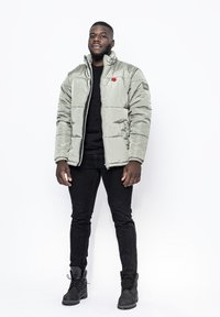 Puffjacka i ljusgrön, med hög krage, dragkedja framtill och en röd logodetalj. Bärs över en svart skjorta och matchas med svarta byxor.