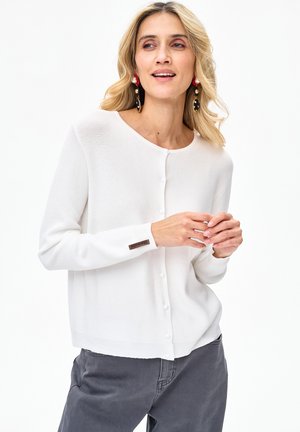 Blondynka w białym, zapinanym na guziki kardiganie, szarych spodniach i dużych kolorowych kolczykach, pozująca na białym tle.