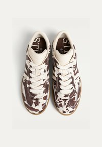 Et par sneakers i hvid og brun kohaleprint med hvide snørebånd og gummisåler, set oppefra på en ensfarvet lys baggrund.
