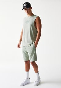 Φωτεινό πράσινο αμάνικο πουκάμισο με εκτυπωμένο λογότυπο, ταιριαστά σορτς, λευκά αθλητικά παπούτσια και μαύρο καπέλο με λογότυπο, σε απλό φόντο.