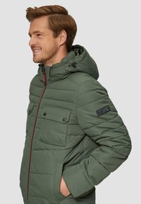 Veste verte rembourrée avec une capuche, fermeture éclair rouge, deux poches poitrine et un patch logo sur la manche. A une texture matelassée.