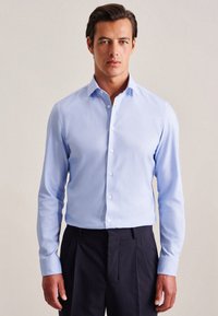 Camicia azzurra a maniche lunghe con colletto classico e bottone frontale, realizzata in tessuto liscio; abbinata a pantaloni scuri.