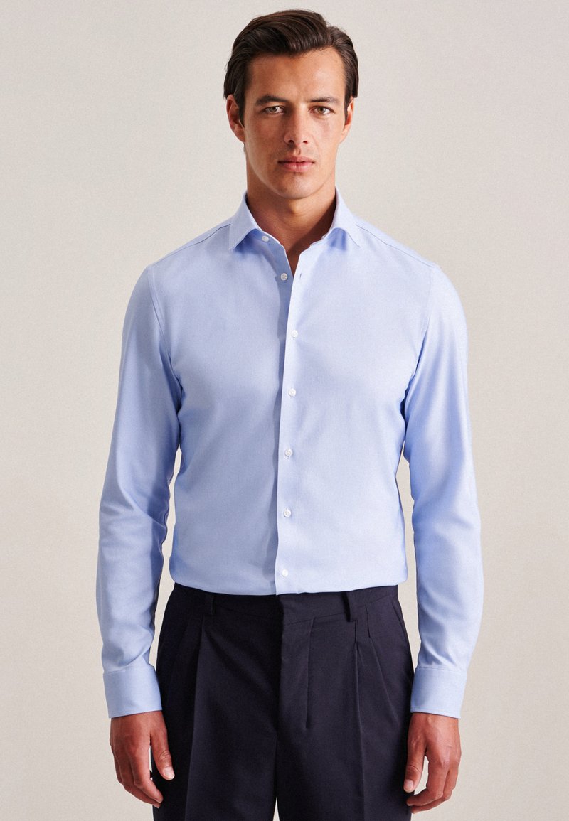 Camicia azzurra a maniche lunghe con colletto classico e bottone frontale, realizzata in tessuto liscio; abbinata a pantaloni scuri.