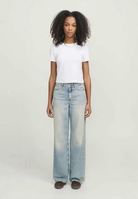 T-shirt branca cropped combinado com jeans de perna larga em azul claro, apresentando um tom desbotado e um leve desgaste. Sapatos castanhos completam o look.