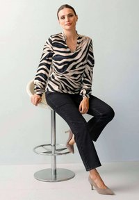 Eine Frau sitzt auf einem weißen modernen Hocker, trägt eine blusenähnliche, gestreifte Zebra-Oberbekleidung, schwarze Hosen und beigefarbene High Heels vor einer schlichten Wand.