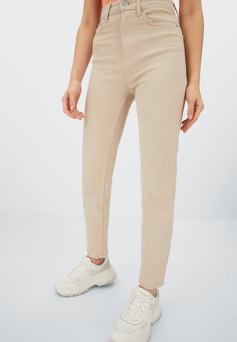 Pantalon beige taille haute et coupe slim porté avec des baskets blanches épaisses et un haut orange, vu de la taille aux pieds.