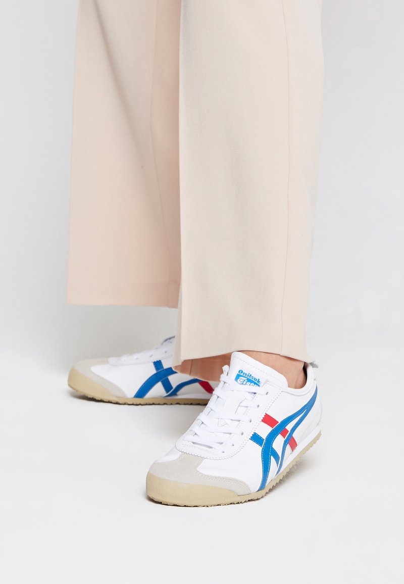 Baskets blanches avec des bandes bleues et rouges portées avec un pantalon beige clair à jambes larges sur un fond clair uni.