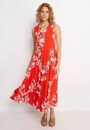 Frau trägt ein ärmelloses rotes Maxikleid mit weißem Blumenmuster und beigen Keilsandalen, steht vor weißem Hintergrund.