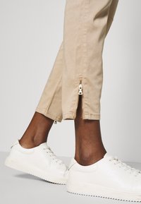 Beige zkrácené kalhoty se bočními zipy a hladkou texturou, spárované s bílými teniskami s texturovanou podešví a minimalistickým designem.