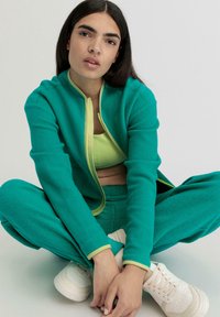 Tuta in pile color turchese con zip e profili giallo brillante. Presenta un crop top aderente sotto. Sneakers leggere completano il look.