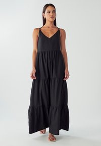 Žena v dlouhé černé maxi šatech s tenkými ramínky, stojící bosky před čistě bílým pozadím.