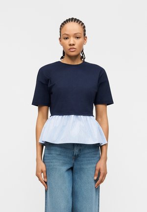 Jeune personne aux cheveux tressés portant un haut à manches courtes bleu marine avec un ourlet à volant rayé bleu clair et un jean en denim à jambes larges.