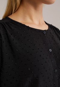 Camicia nera con bottoni, con motivo a punti a rilievo, scollatura rotonda e maniche corte. Presenta due bottoni frontali per la chiusura.
