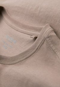 Beige bavlněné tričko s límcem hladké textury. Má žebrovaný výstřih a malou cedulku s modrým textem, která uvádí značku a materiál.