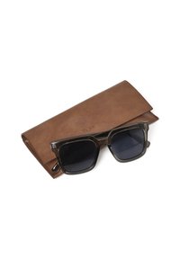 Des lunettes de soleil avec une monture gris foncé et des verres noirs, posées sur un étui en cuir marron avec une texture lisse et un logo en relief.