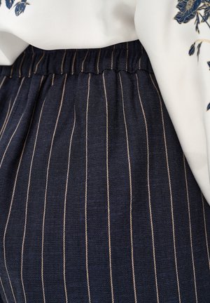 Donkerblauwe broek met dunne beige verticale krijtstrepen en een elastische tailleband, gecombineerd met een witte stof met blauwe bloemenborduursels.