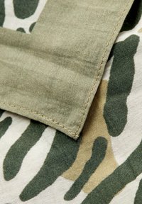 Camouflage stof med en blanding af grønne og beige mønstre, med en glat tekstur. En foldet kant med en syet søm er synlig.