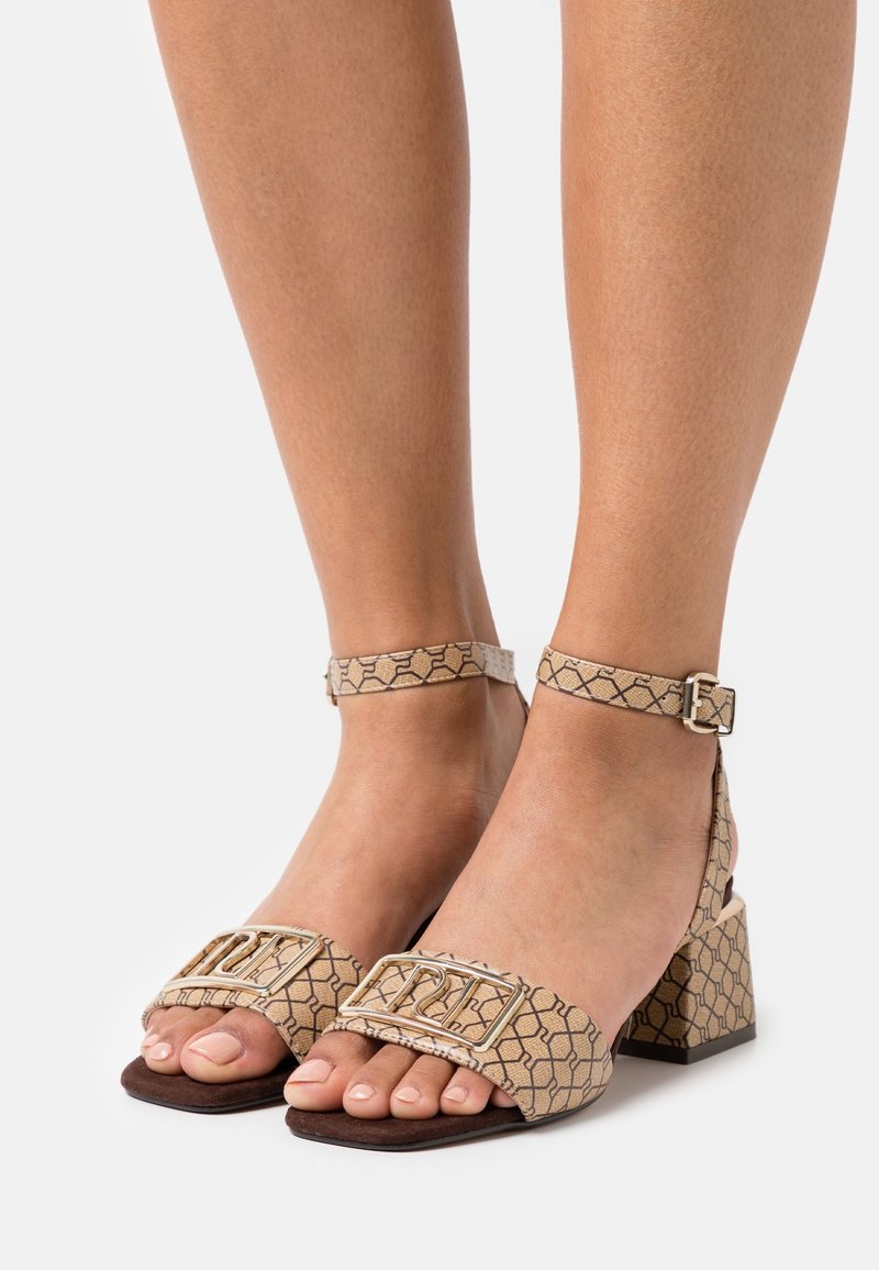 Pieds portant des sandales à talons blocs beige avec des brides à la cheville et un motif géométrique sur un fond blanc.