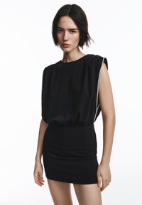 Črna obleka z ohlapnim, preloženim zgornjim delom in oprijeto krilom. Mehak material z elastičnim pasom, brezrokavnik in prefinjenimi šivi.
