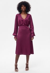 Satynowa sukienka midi w głębokim bordowym kolorze, z długimi rękawami, głębokim dekoltem w serek i marszczonym pasem. Gładka faktura z płynną sylwetką.