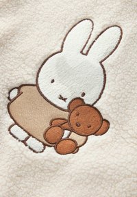 Kreemivärvi flanellkangas, millel on broderitud jänku, kes hoiab käes pruunist teddy-karu, kaunistatud heleda pruunide ja valgete detailidega. Pehme tekstuur.