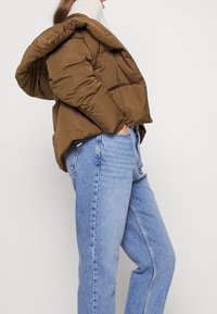 Chaqueta acolchada marrón con capucha, que presenta un acabado mate, cierre frontal de botones y costuras diagonales. Combinada con pantalones vaqueros de pierna recta de color azul claro.
