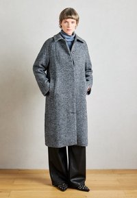 Pelēks vilnas herringbone mētelis ar apkakli, vienu pogu un sānu kabatām; apvienots ar melnām ādas platām biksēm un smailiem apaviem.