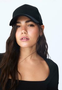 Crna korduroy kapa s zakrivljenim vizirom, glatkom teksturom i detaljima šivanja. Nosila je osoba s dugom, tamnom kosom i crnom majicom.