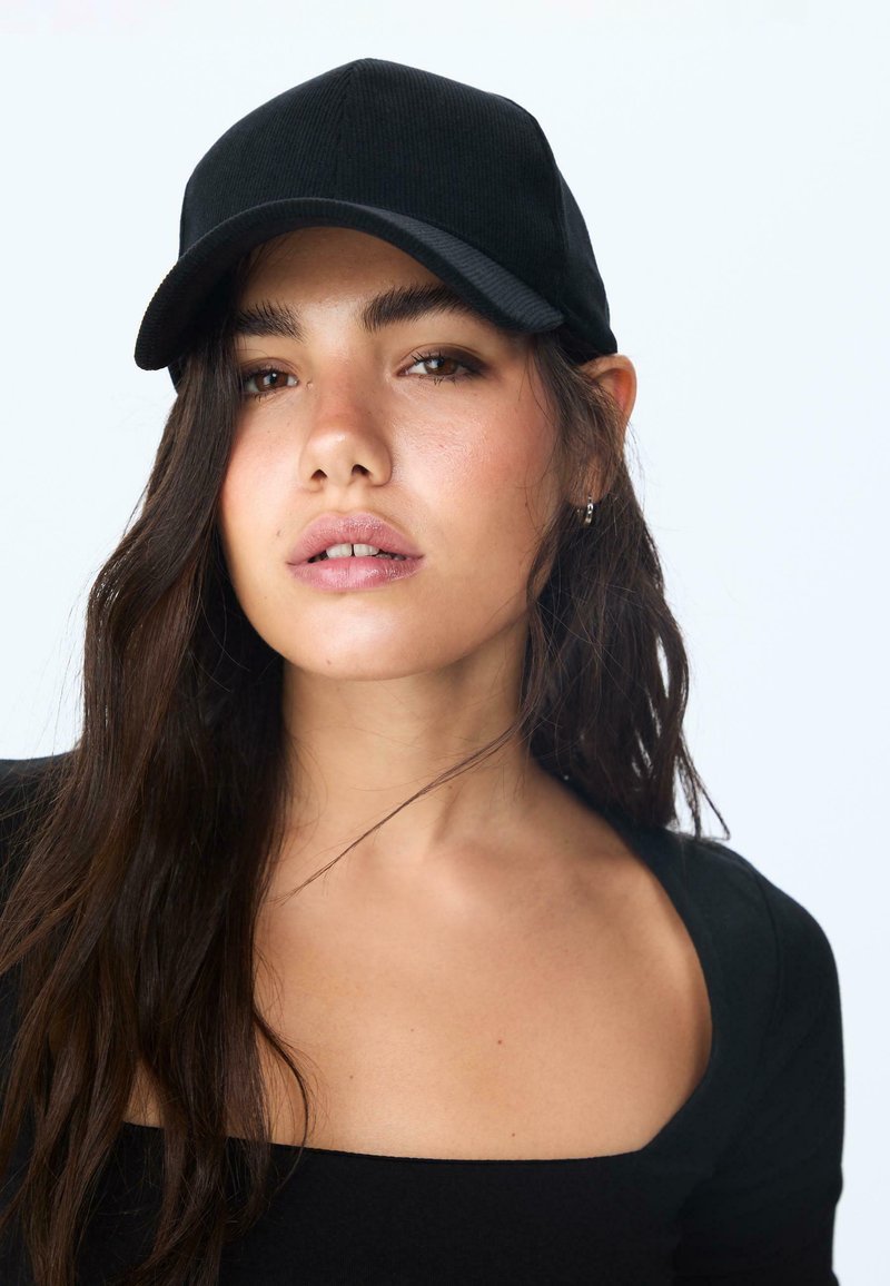 Crna korduroy kapa s zakrivljenim vizirom, glatkom teksturom i detaljima šivanja. Nosila je osoba s dugom, tamnom kosom i crnom majicom.