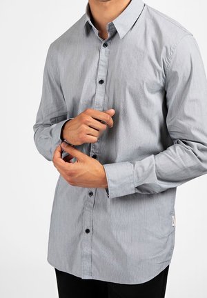 Mężczyzna poprawia mankiet jasnoszarej, długorękawowej koszuli z czarnymi guzikami, stojąc na jednolitym białym tle.