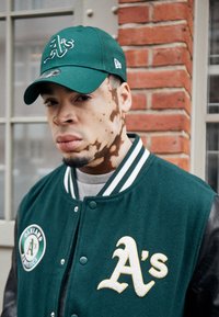 Πράσινη καμπάνα και μπουφάν των Oakland Athletics, με χνουδωτή υφή, λευκό λογότυπο και αντίθετα δερμάτινα μανίκια σε τοίχο από τούβλα.