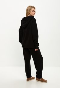 Černá kapucínová mikina s žebrovanými okraji, spárovaná s černými kalhotami volného střihu a hnědými slip-on botami. Jednoduchý, jednobarevný design.