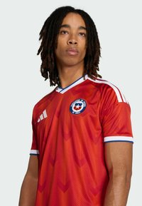 Młody mężczyzna z dredami w czerwonej koszulce reprezentacji Chile w piłce nożnej z białymi i niebieskimi wykończeniami, patrzący lekko w górę.