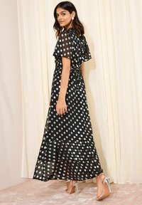 Langes schwarzes Kleid mit silbernen Punkten, transparenter Stoff, schimmernder Saum, kurze, flatternde Ärmel und taillierter Schnitt, kombiniert mit metallischen Absatzschuhen.