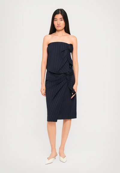 Vrouw in een strapless marineblauwe pinstriped jurk met ruches, gecombineerd met witte puntige kittenheels, staand op een effen achtergrond.