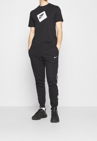 Černé tričko s bílou grafikou "JUMP MAN", kombinované s černými vzorovanými tepláky s logem Nike. Stylizováno s černými sportovními botami.