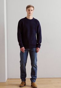 Marine gebreide trui met ronde hals, lange mouwen en ribgebreide manchetten, gecombineerd met blauwe jeans en bruine laarzen op een houten vloer.