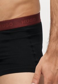 Torse et main masculins portant un boxer noir avec une ceinture bordeaux siglée "HUGO" sur fond blanc.