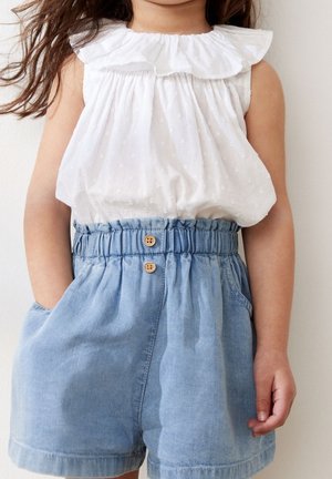Enfant portant un chemisier blanc sans manches avec un col à volants et un short taille haute bleu clair avec une ceinture élastique et des boutons en bois.